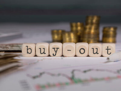 The words buy-out in wooden blocks on top of graphs and charts with stacks of money.